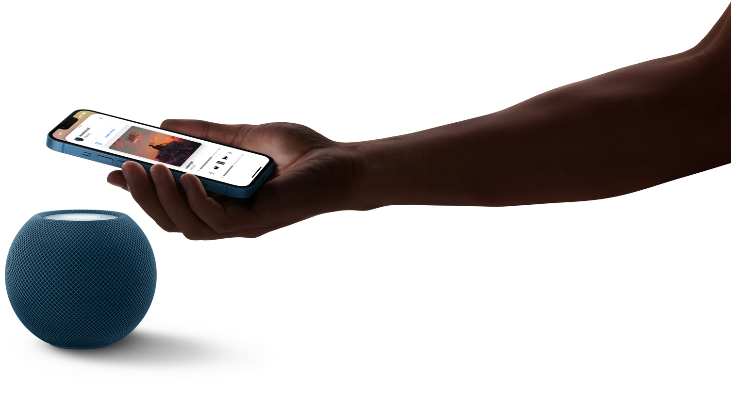 Blue HomePod mini with a person&rsquo;s hand holding an iPhone over HomePod mini. Music is playing on the iPhone screen.