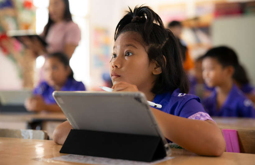 Uma menina está sentada em sua mesa, olhando com atenção para a frente da sala de aula. Ela segura um Apple Pencil sobre um iPad