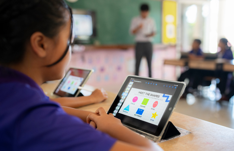 Close de uma aluna na sua mesa estudando em um iPad. A tela exibe formas coloridas com o título 
