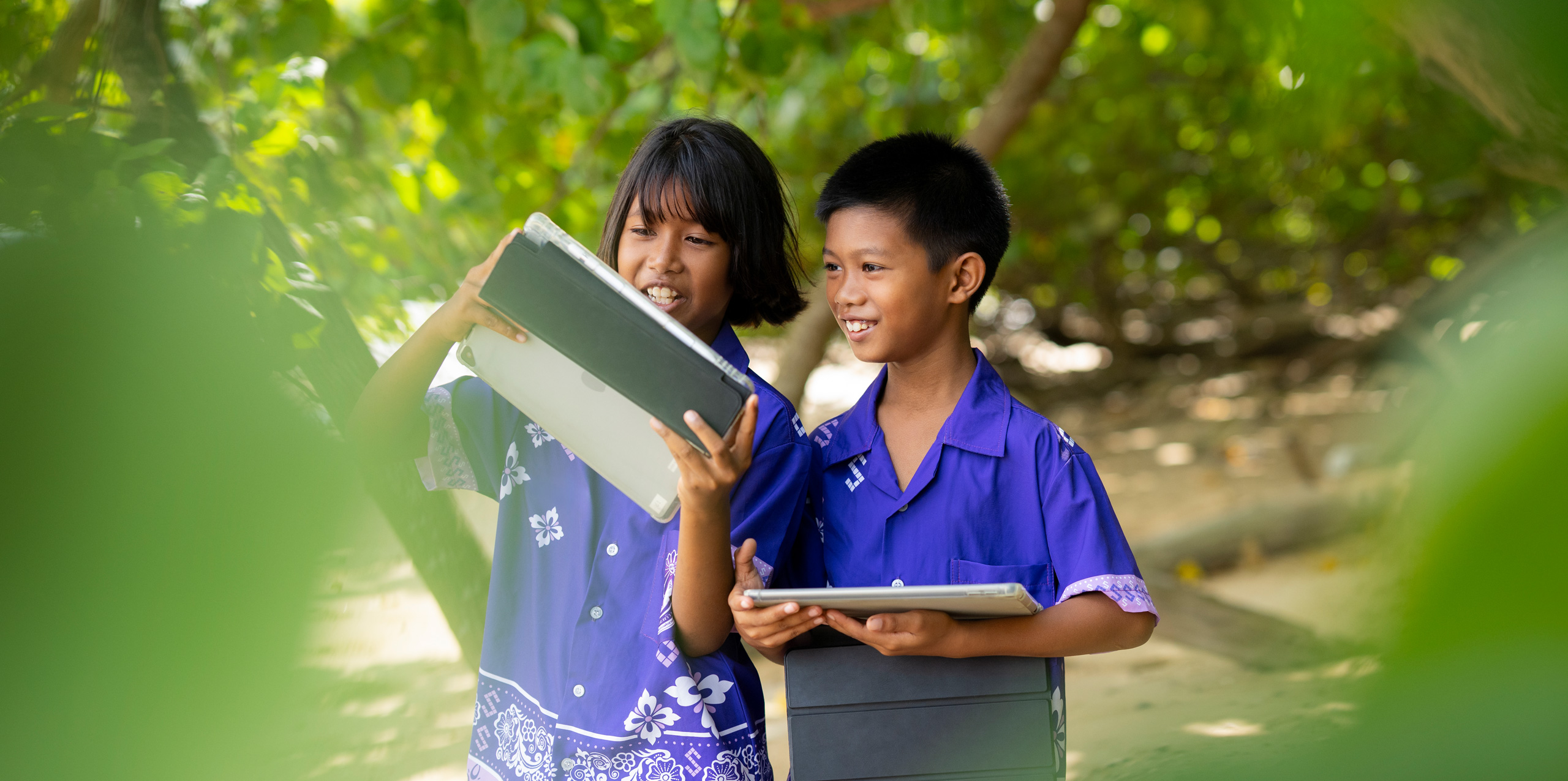 Duas crianças do ensino fundamental, uma menina e um menino, estão em pé ao ar livre cercadas de árvores tropicais, cada uma segurando um iPad As duas estão olhando para a tela do iPad da menina, sorrindo e atentas