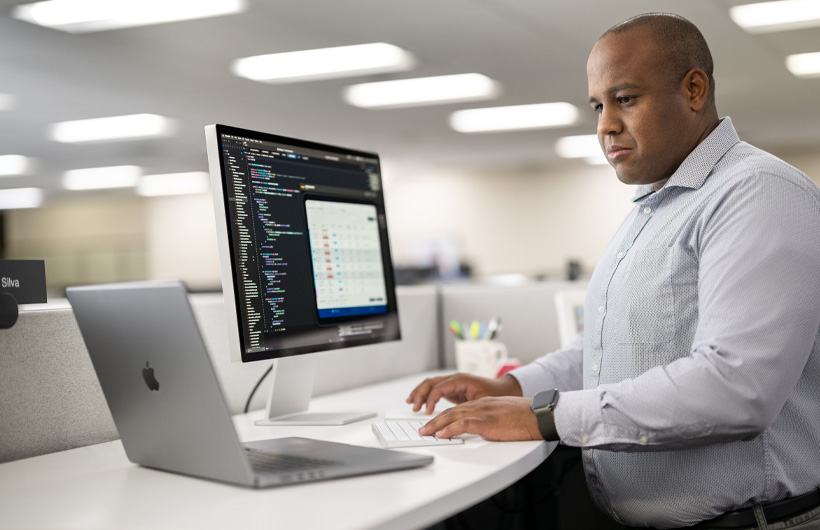 En utvikler står ved arbeidsbordet sitt og bruker en Mac og MacBook. En kode og appinformasjon vises på Mac-skjermen