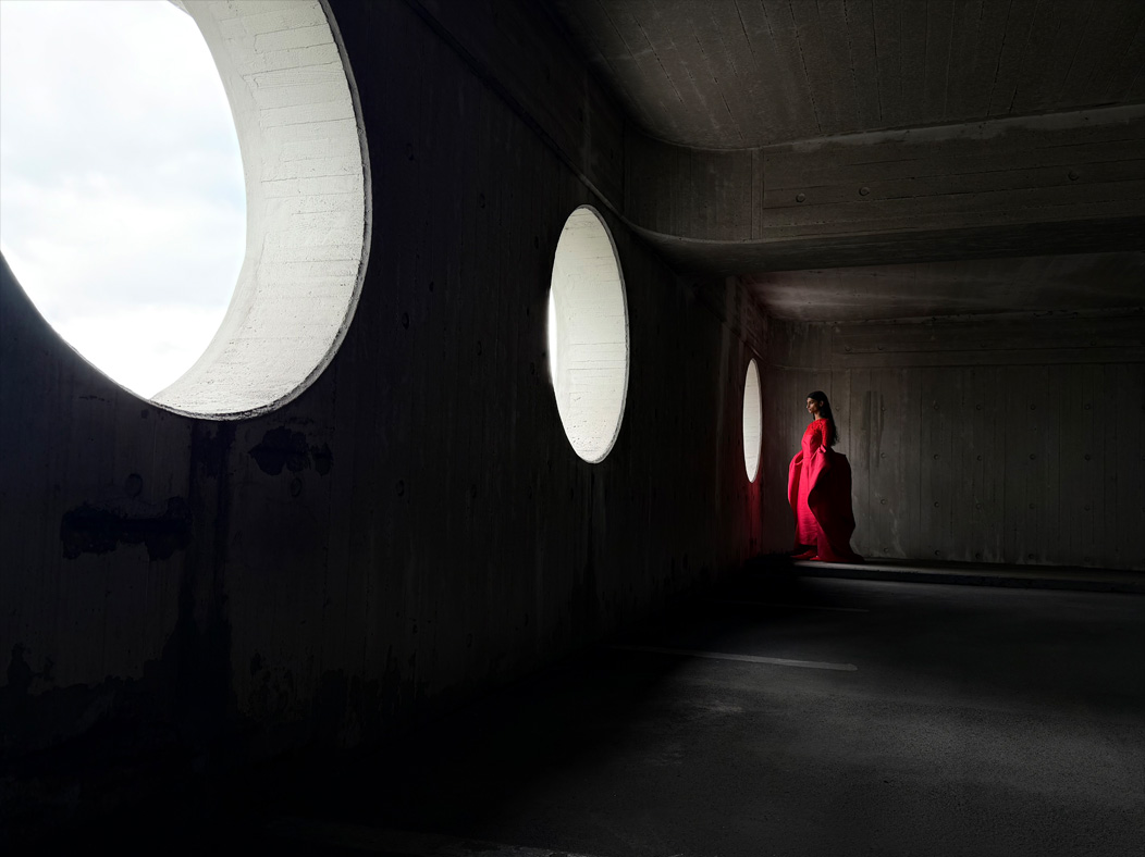 En kvinde i en rød kjole står i et tomt rum og bliver fotograferet. Fotos-appen skifter mellem brændvidder for at demonstrere mulighederne i Pro-kamerasystemet og viser: 28 mm